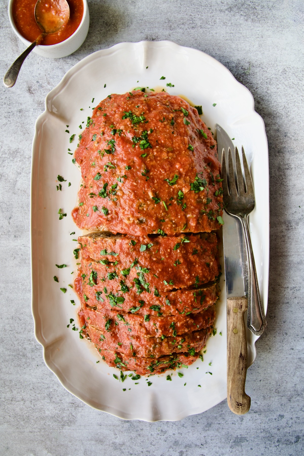 Italian Meatloaf on white platter with serving utensils garnished with chopped fresh parsley.