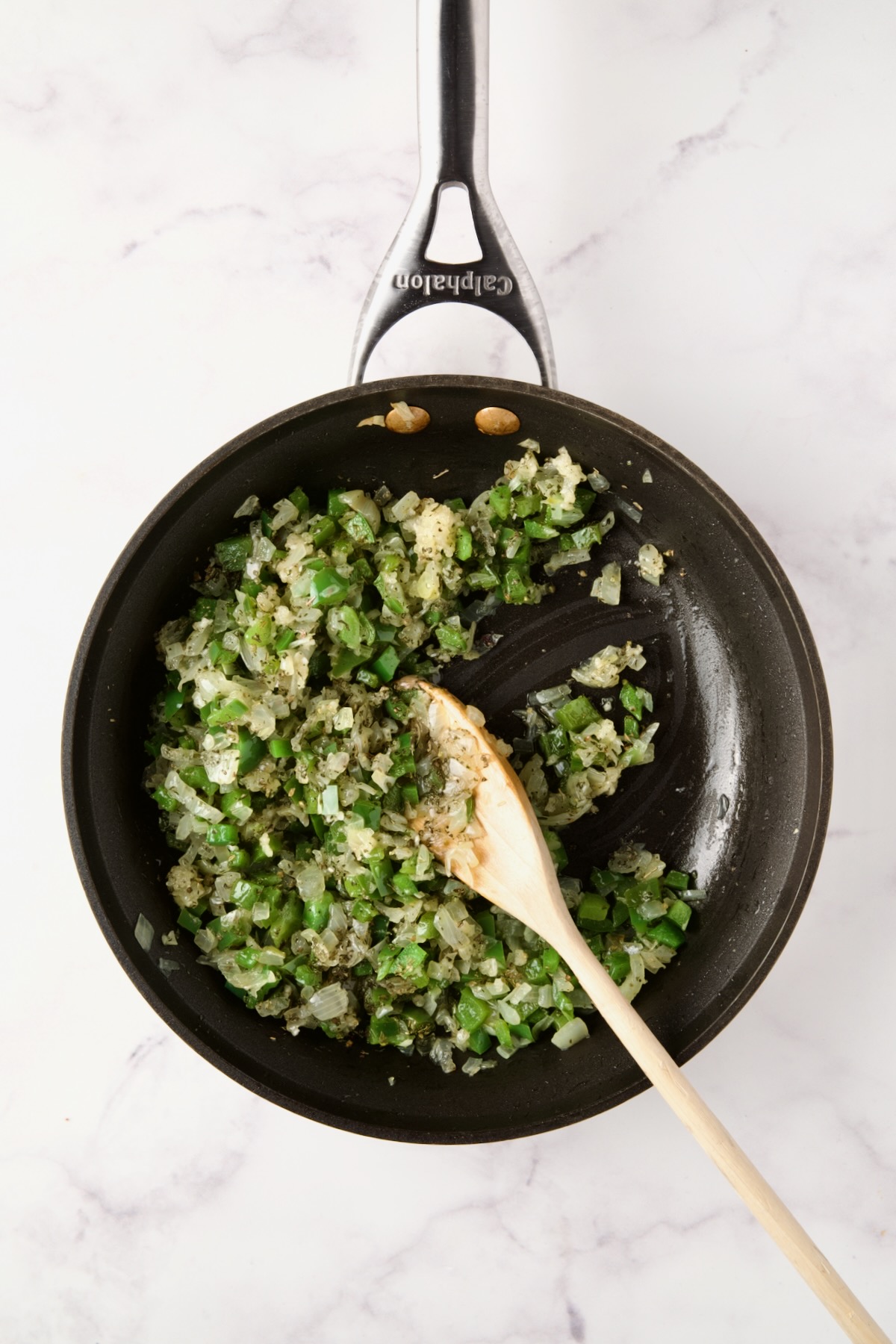 Cooked onions and green pepper in nonstick skillet with white spoon stirring in dried herbs and garlic.