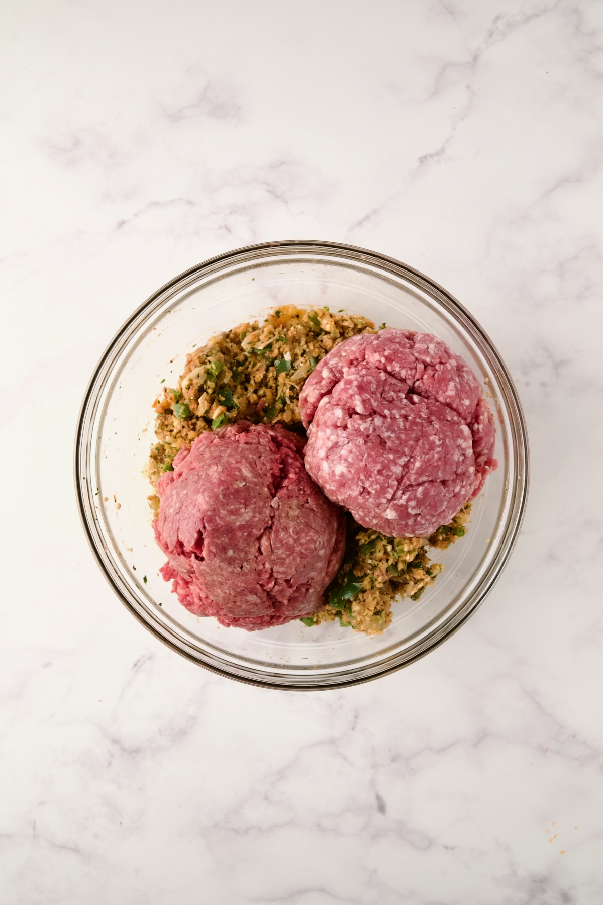 Ground meats added to other ingredients in glass bowl.