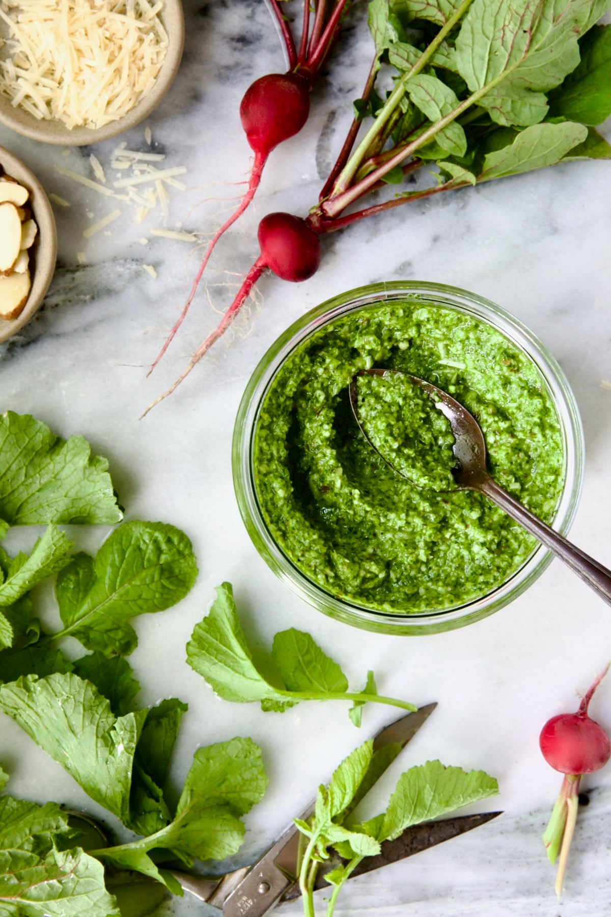Radish Greens Pesto in glass jar with spoon on marble surface with ingredients in the pesto around it.