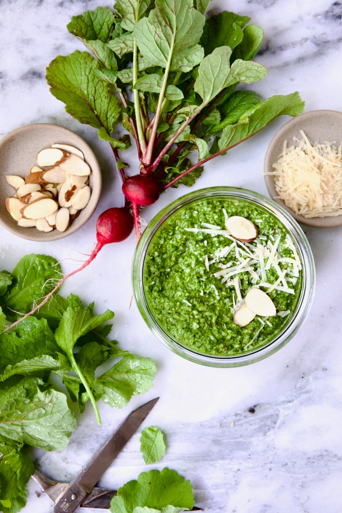 Radish Greens Pesto in glass jar with spoon on marble surface with ingredients in the pesto around it.