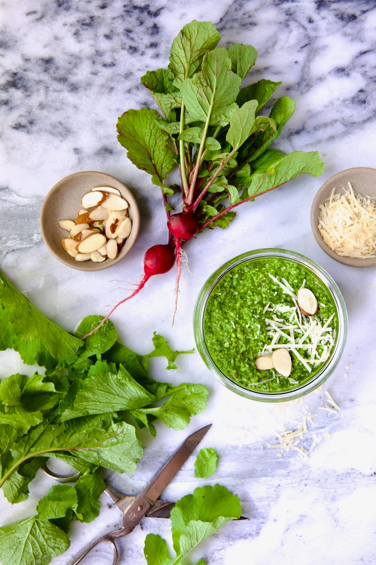 Radish Greens Pesto in glass jar with spoon on marble surface with ingredients in the pesto around it.