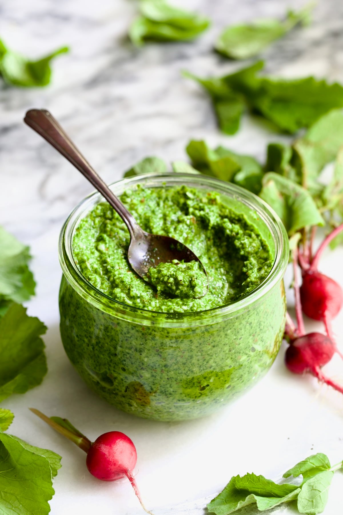 Radish Greens Pesto in glass jar with radish greens scattered around it.