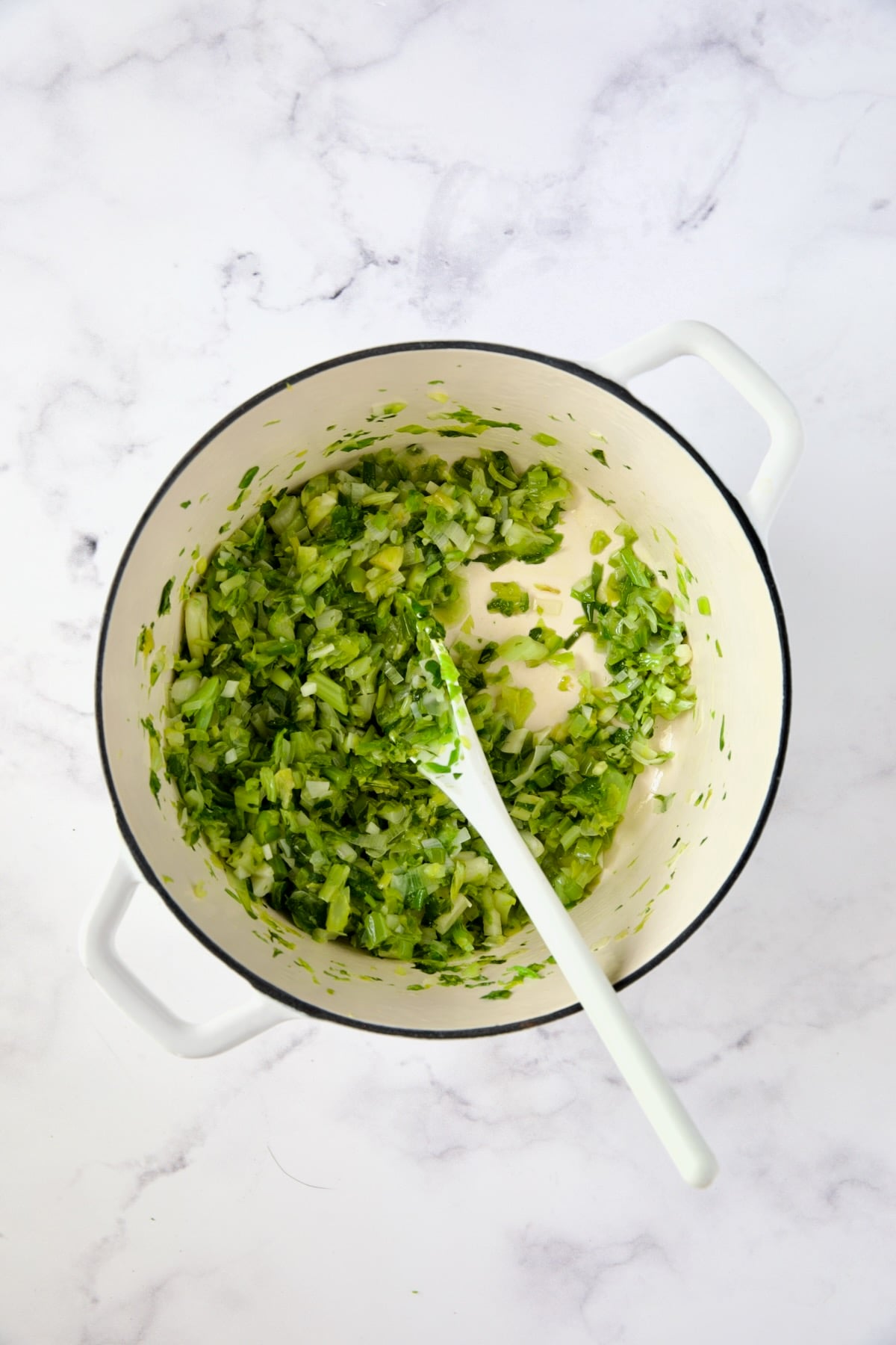 Scallion and celery leaves being sauteed in white Dutch oven with white mixing spoon.
