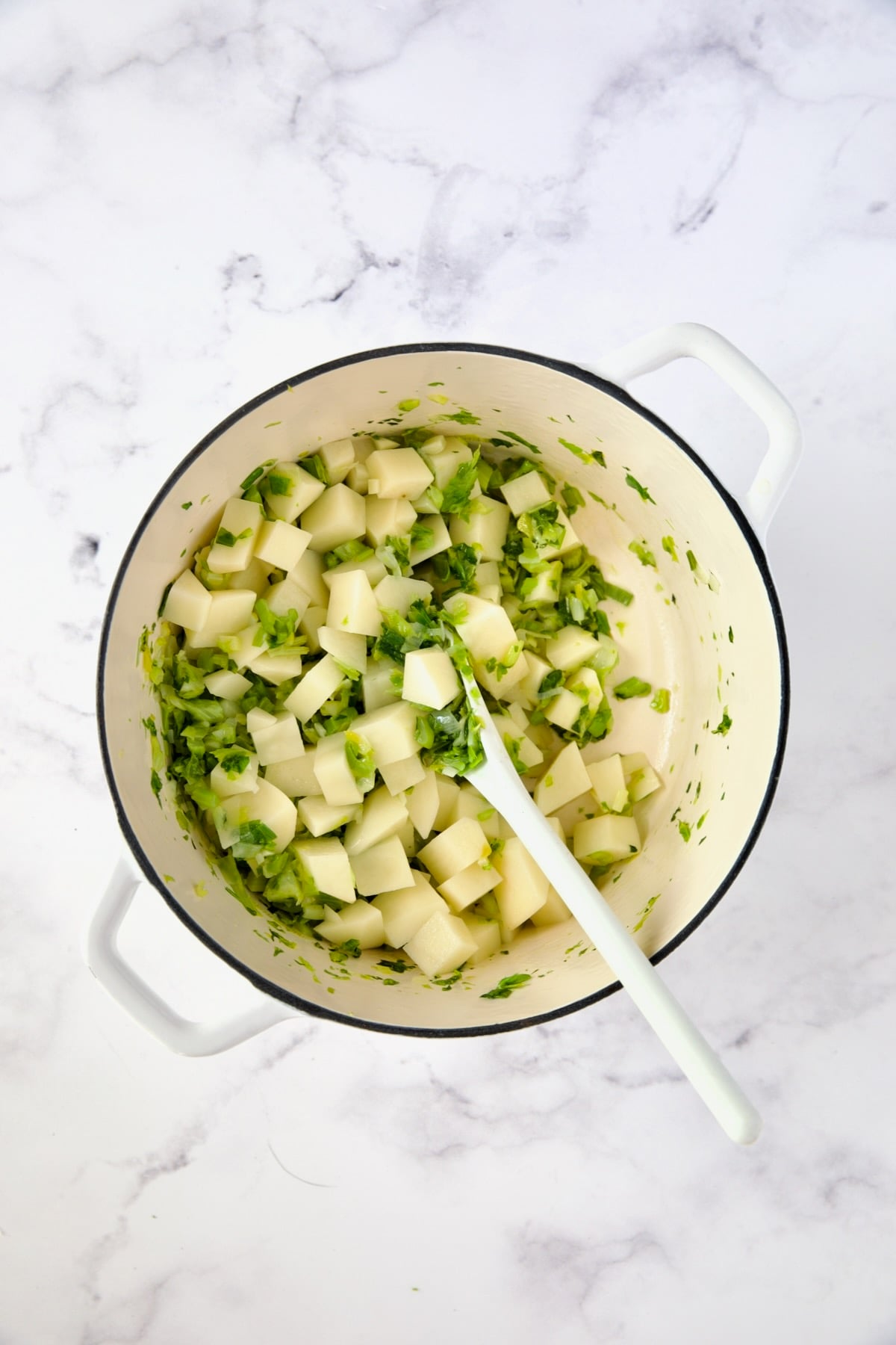 Potatoes added to scallions and celery leaves in white Dutch oven.