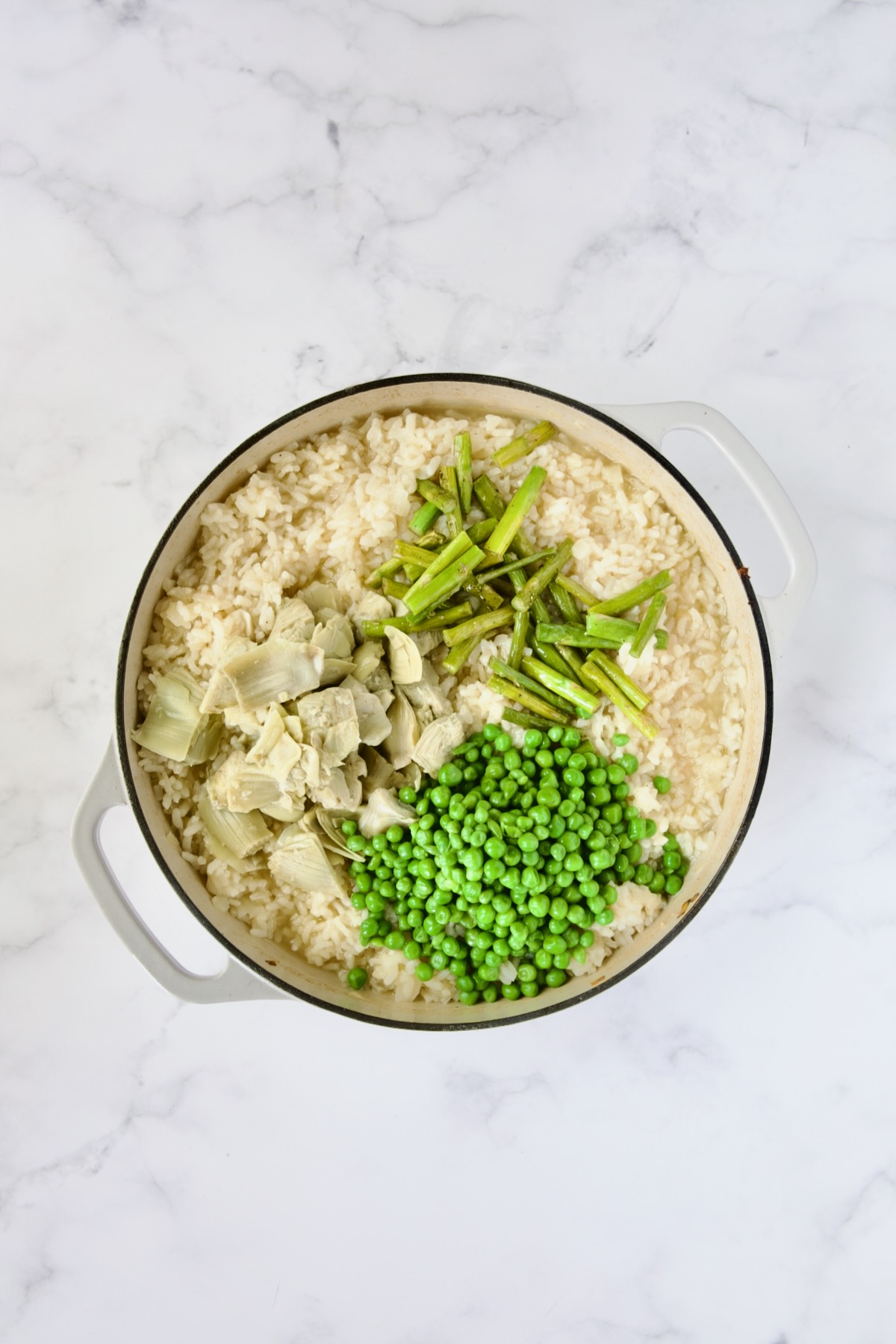Asparagus stems, peas and artichoke hearts added to cooked risotto in white enamel cast iron skillet.