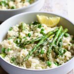 One bowl of Easy Risotto Recipe with Spring Vegetables with asparagus spears on the top and lemon wedge on whitewashed cutting board.