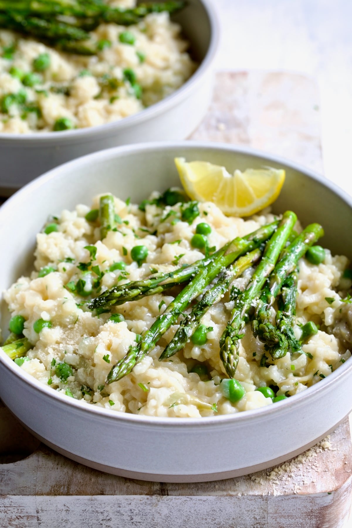 One bowl of Easy Risotto Recipe with Spring Vegetables with asparagus spears on the top and lemon wedge on whitewashed cutting board.