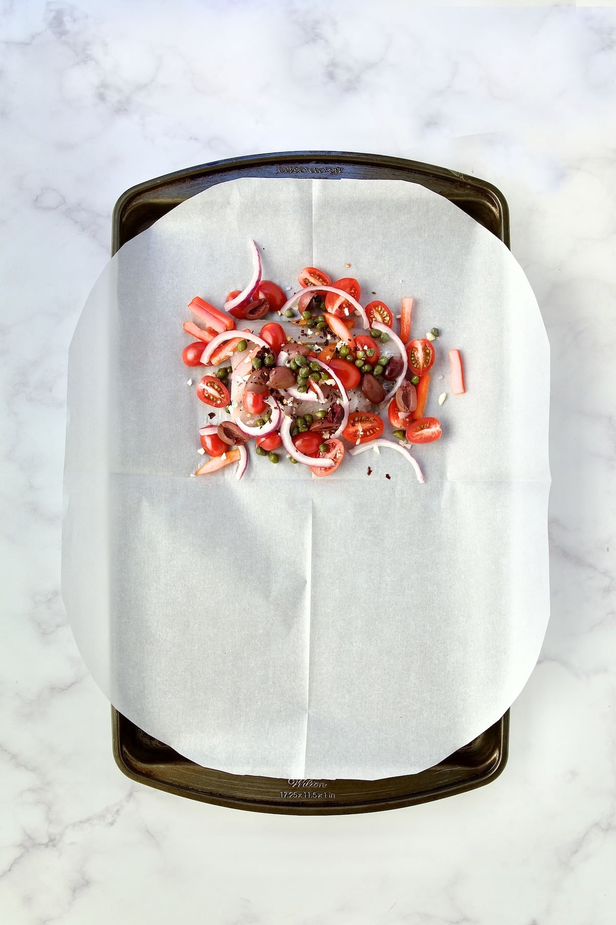 Uncooked fish with other ingredients on parchment paper on rimmed sheet pan.