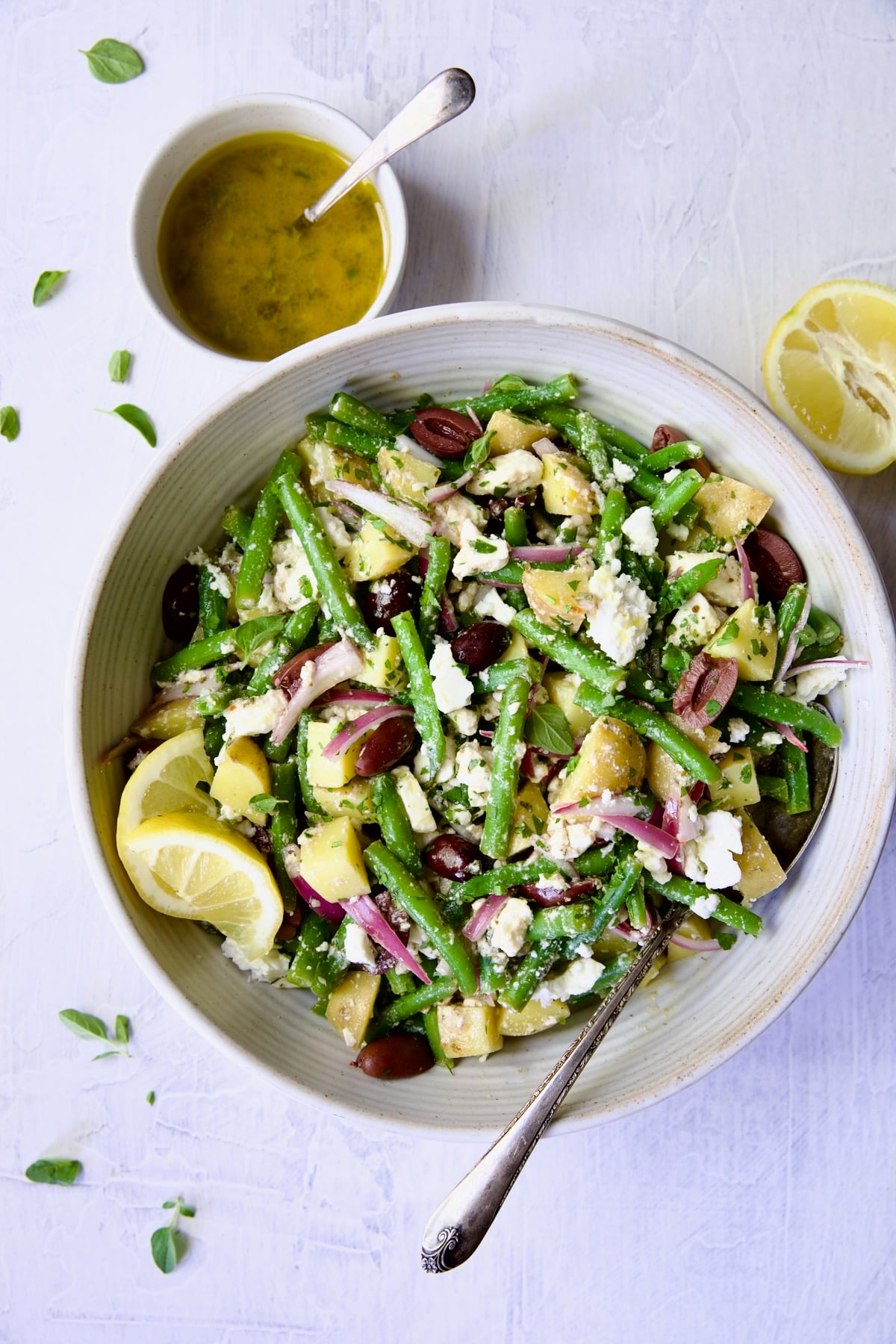 Potato Green Bean Salad in white bowl with serving spoon, vinaigrette and cut lemon.