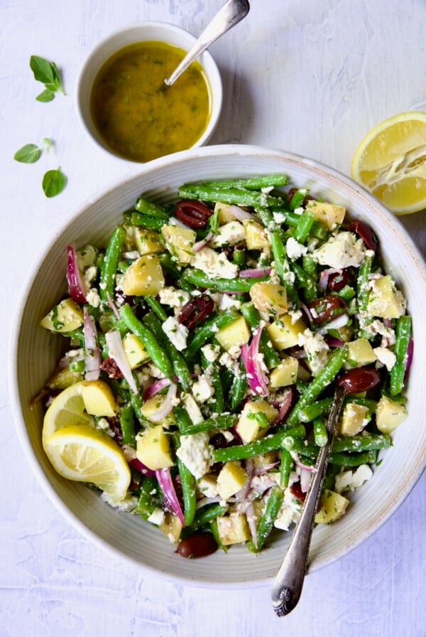 Potato Green Bean Salad in white bowl with serving spoon, dressing and cut lemon.