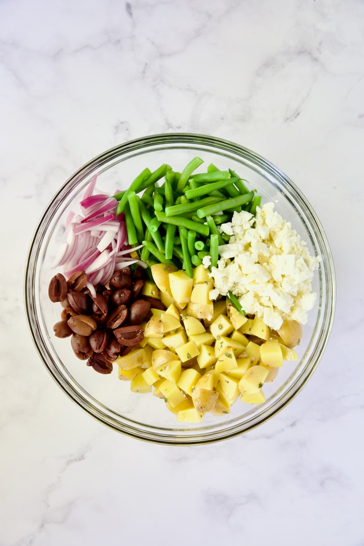 Red onion, blanched green beans, feta cheese, cooked Yukon Gold potatoes and black olives in glass bowl before being stirred together.