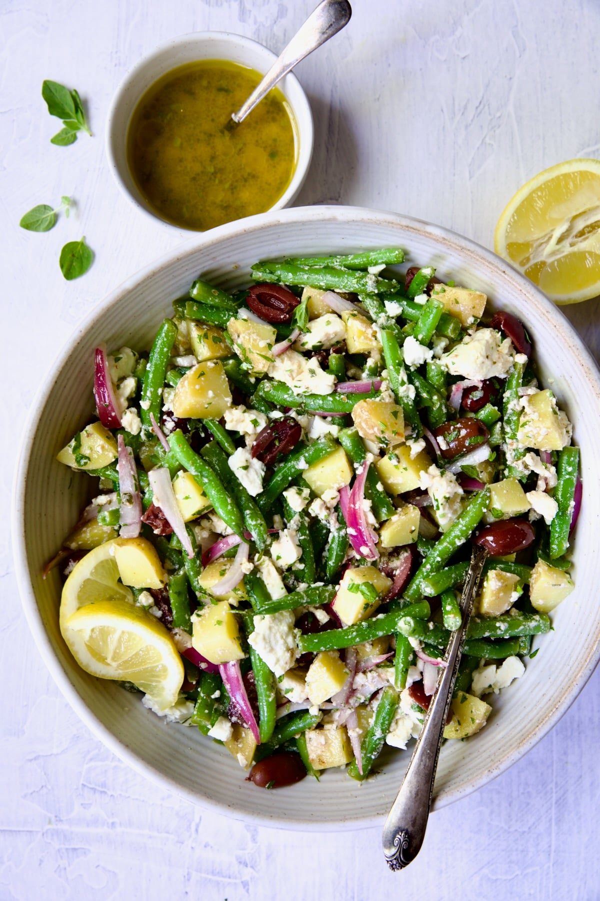 Potato Green Bean Salad in white bowl with serving spoon, dressing and cut lemon.
