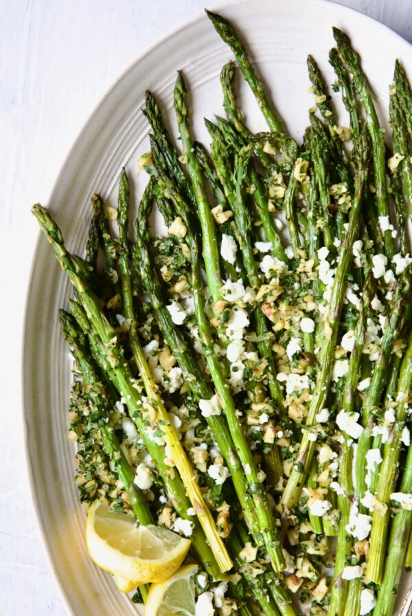 Roasted Asparagus with Walnut Lemon Gremolata and Goat Cheese on white oval platter with lemon wedges.