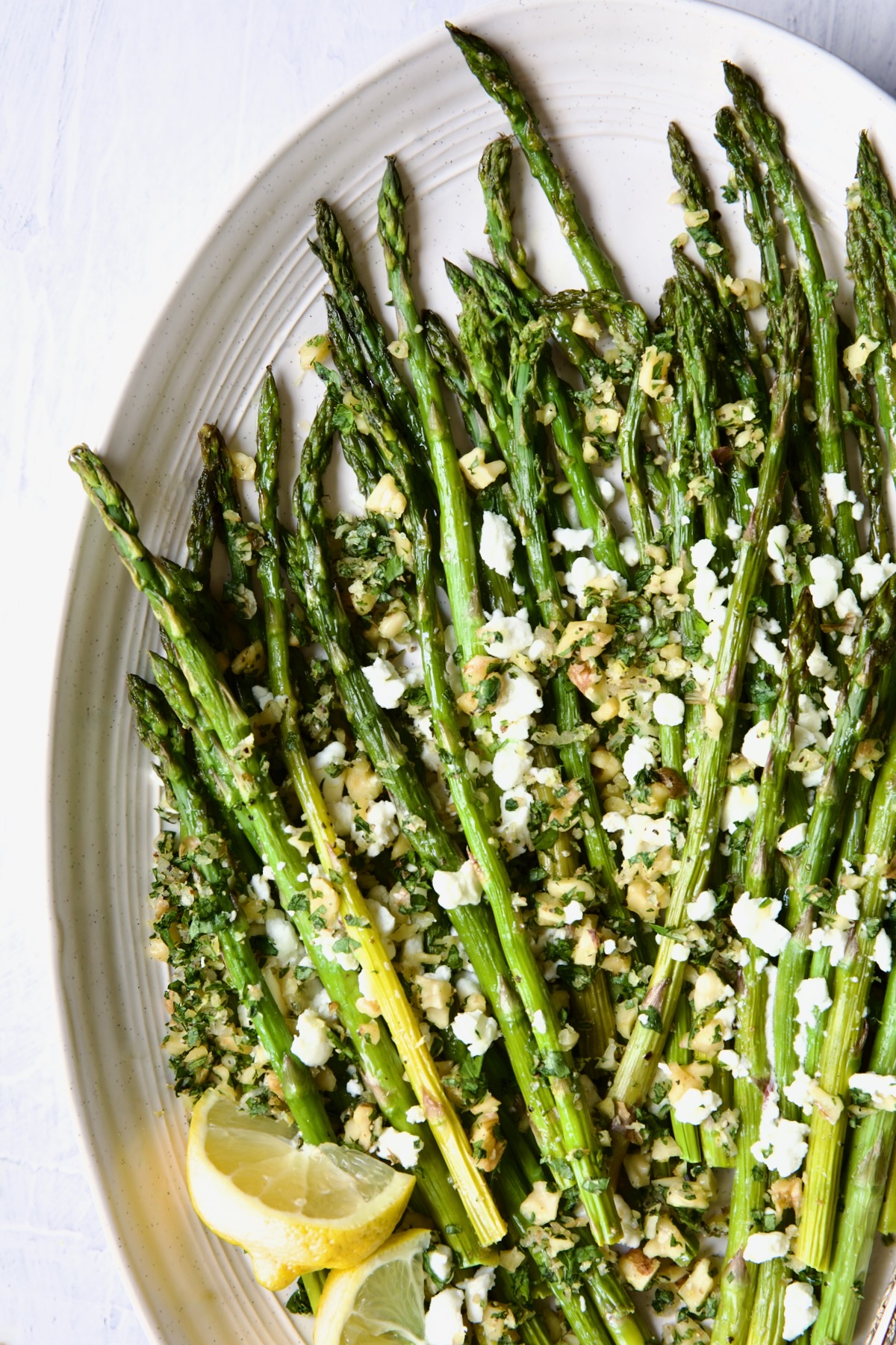 Roasted Asparagus with Walnut Lemon Gremolata and Goat Cheese on white oval platter with lemon wedges.