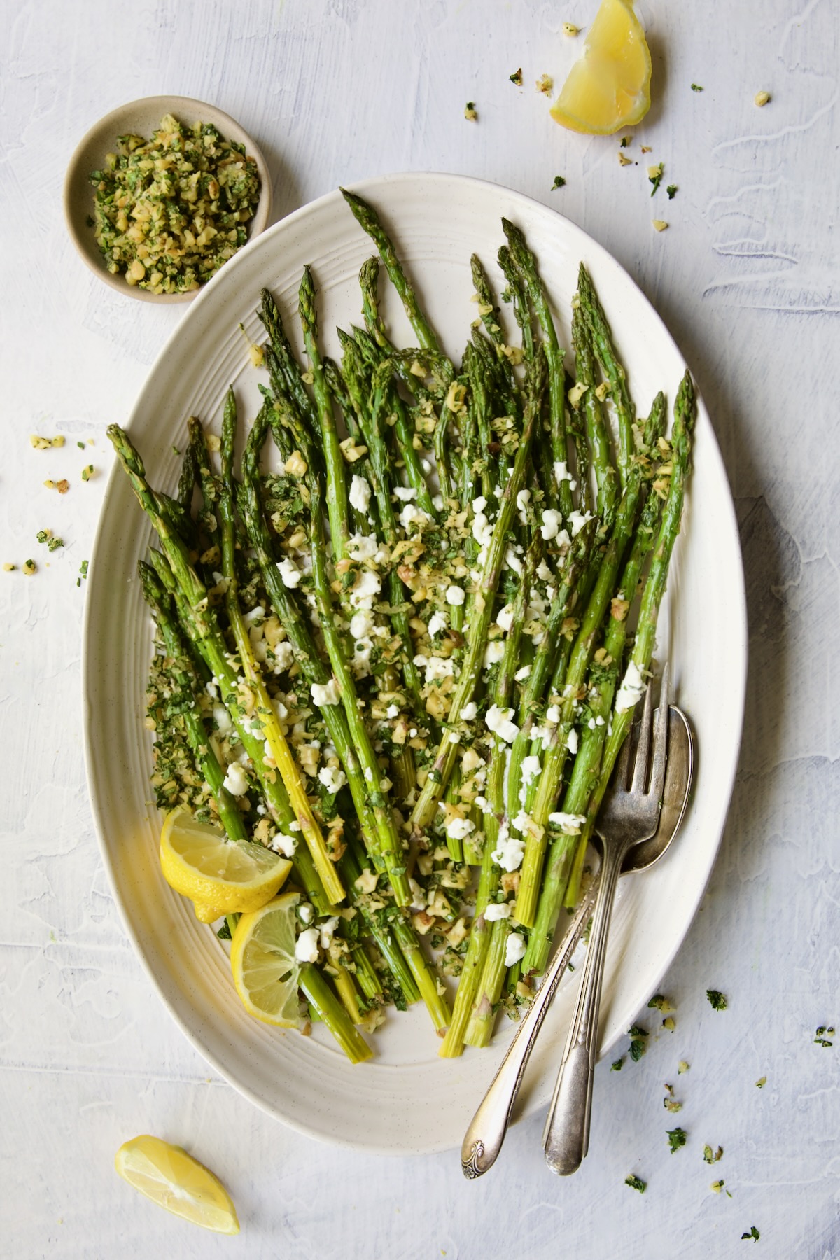 Roasted Asparagus with Walnut Lemon Gremolata and Goat Cheese with lemon wedges, serving utensils and bowl of gremolata.