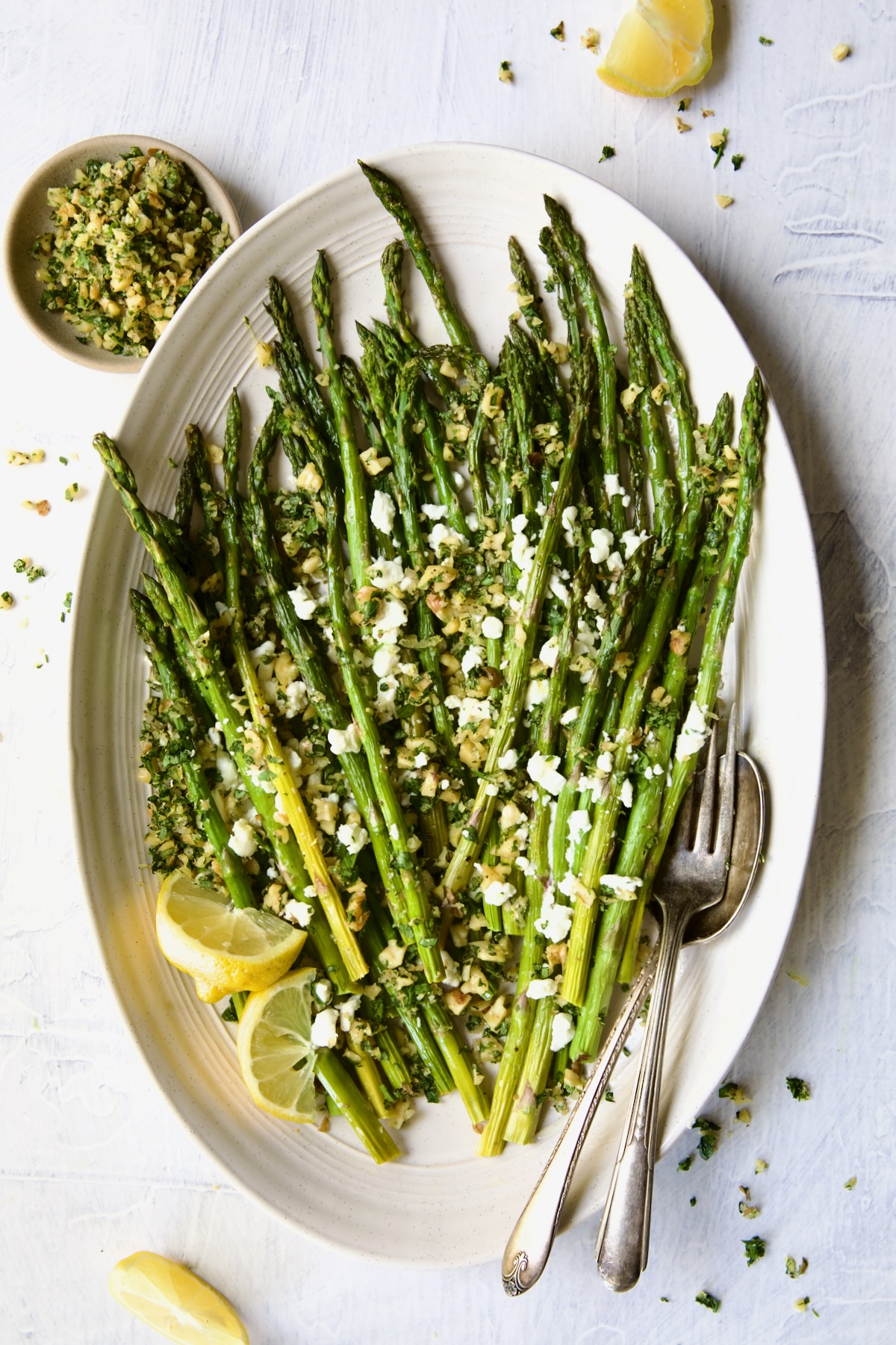 Roasted Asparagus with Walnut Lemon Gremolata and Goat Cheese on oval white platter with lemon wedges and serving utensils.