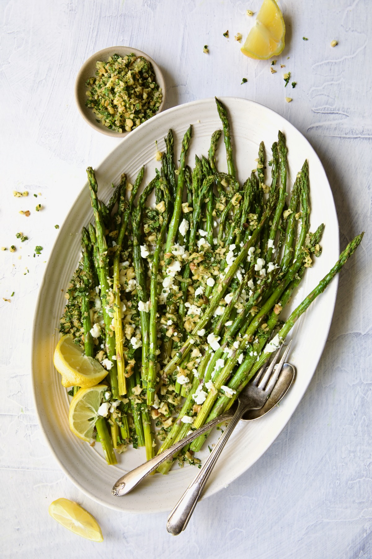Roasted Asparagus with Walnut Lemon Gremolata and Goat Cheese on white oval platter on an angle with lemon wedges, serving utensils and bowl of gremolata.