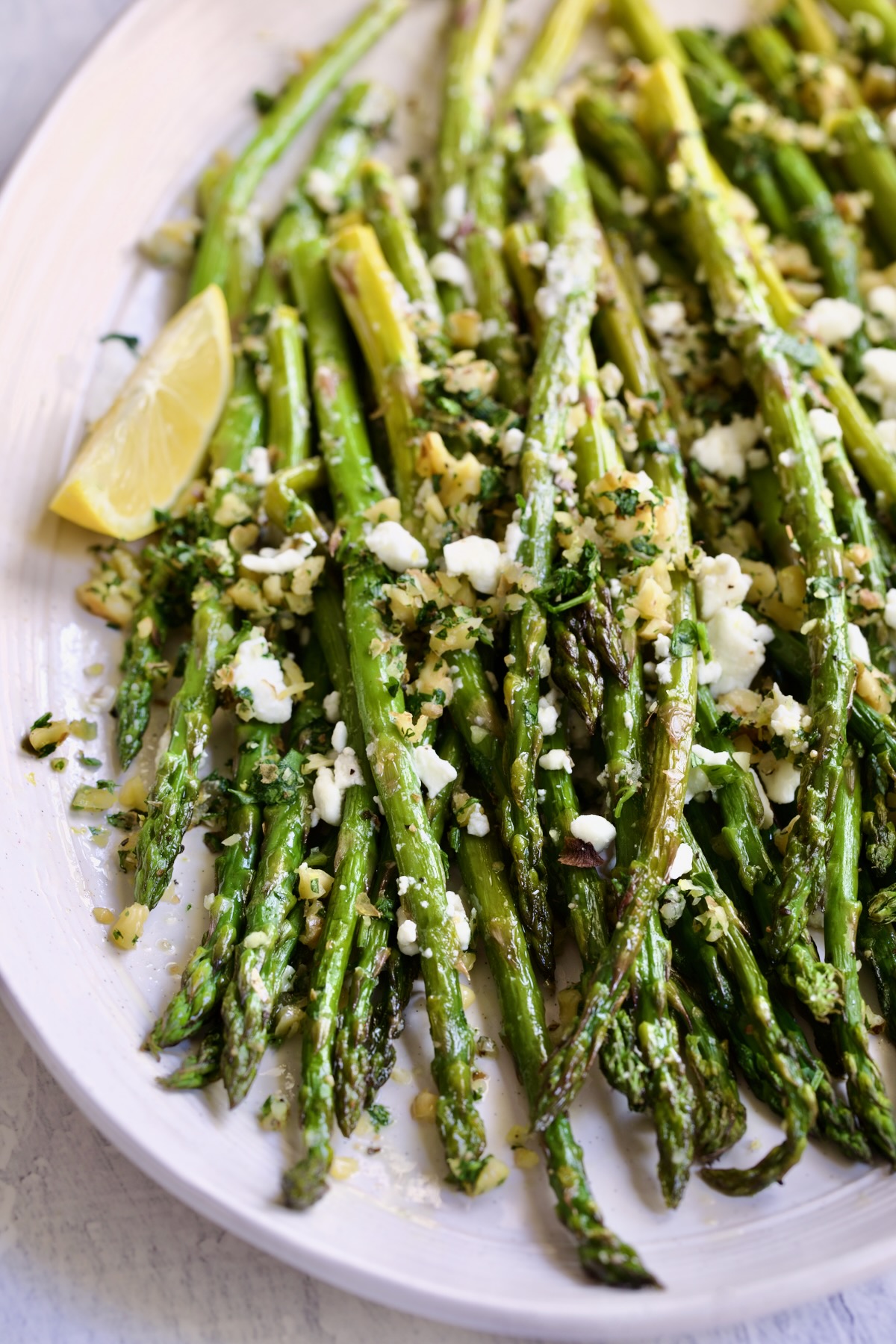 Roasted Asparagus with Walnut Lemon Gremolata and Goat Cheese on white oval platter with lemon wedge.