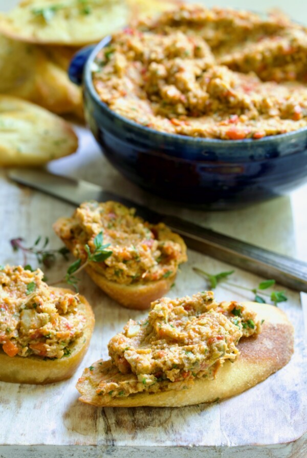 Three pieces of crostini topped with Artichoke Tapenade with bowl of tapenade in the background.