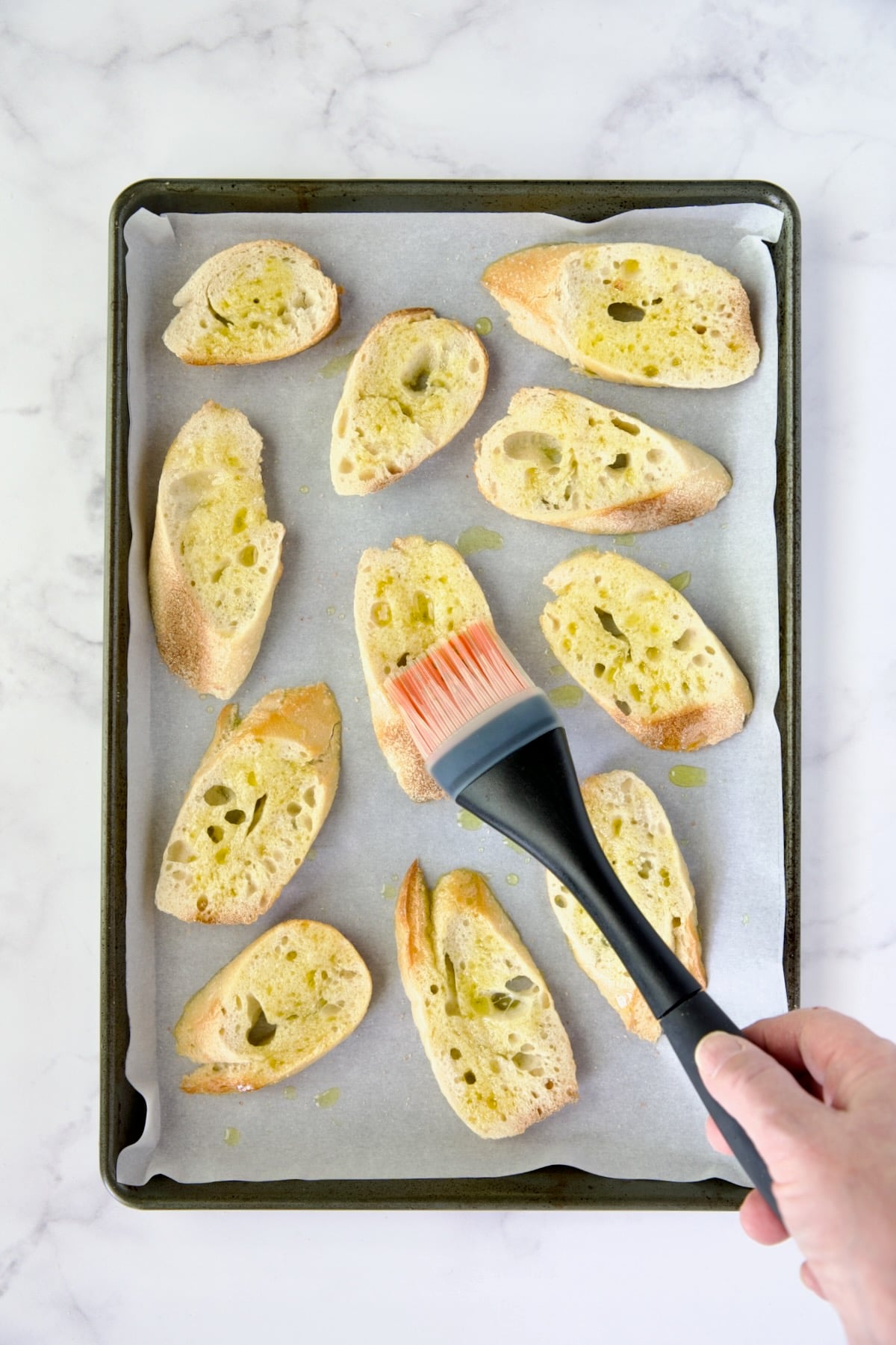 Slices of French baguette being brushed with olive oil on parchment paper-lined baking sheet.
