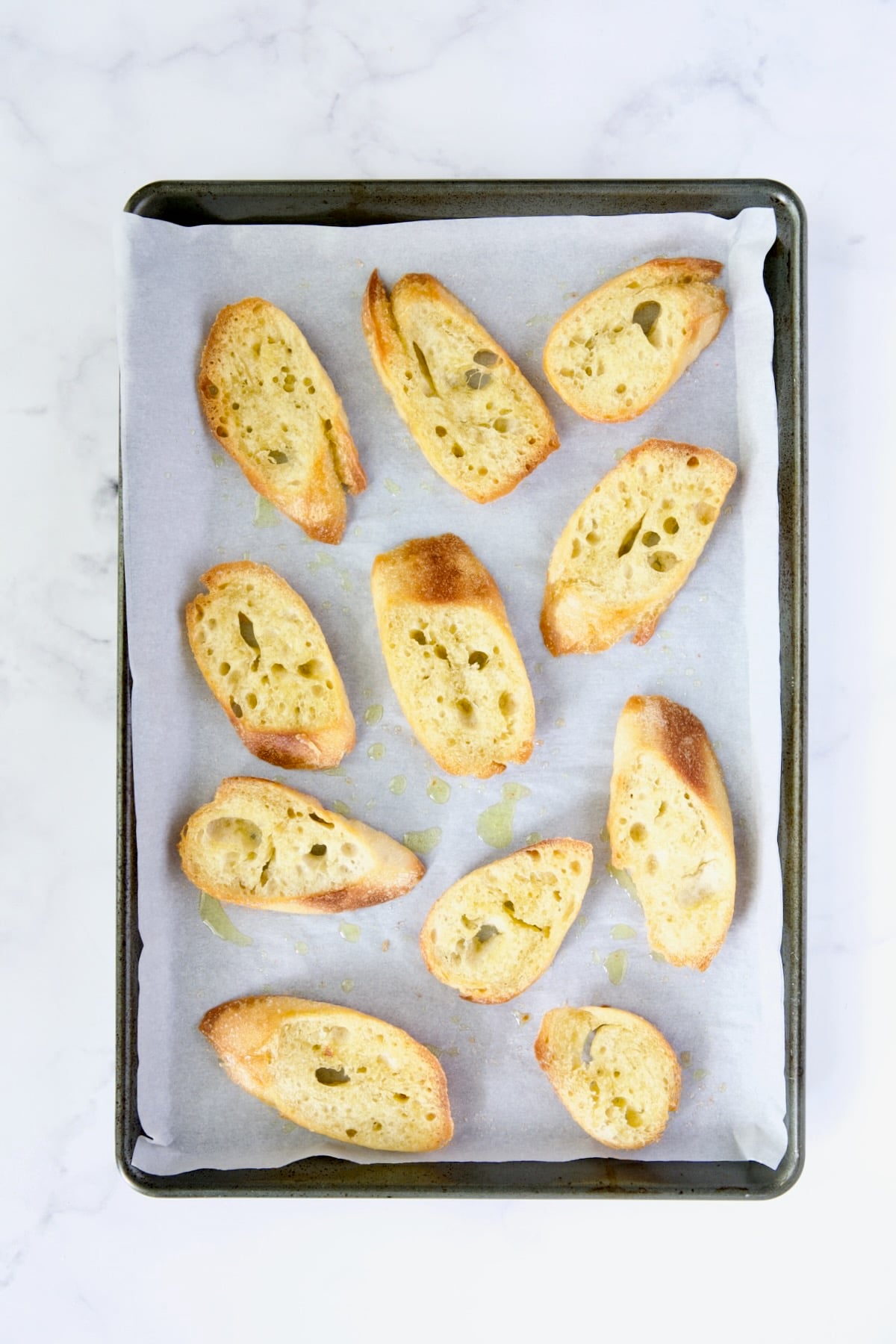 Eleven pieces of fresh bread brushed with olive oil on parchment paper-lined rimmed baking sheet.