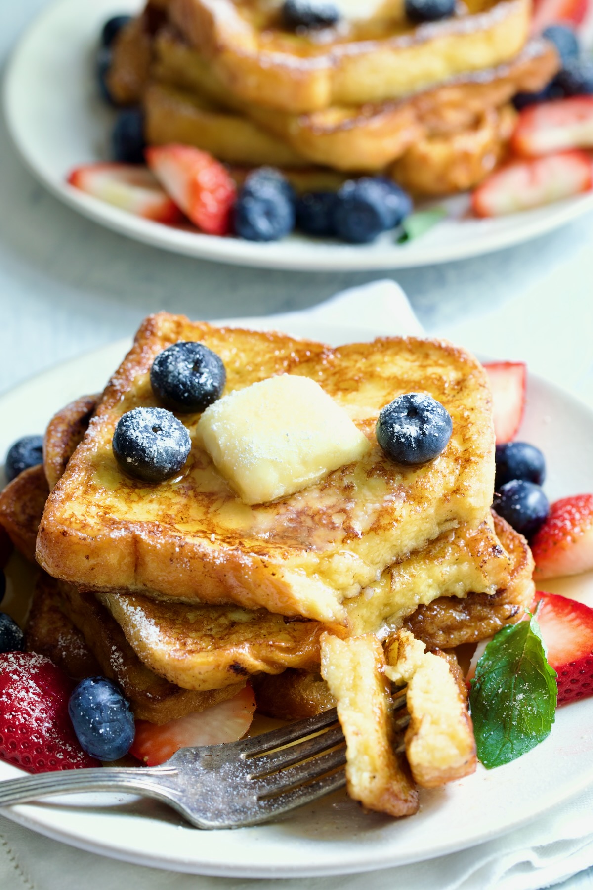 Brioche French Toast on white plate with pat of butter, blueberries and strawberries with two pieces on a fork.