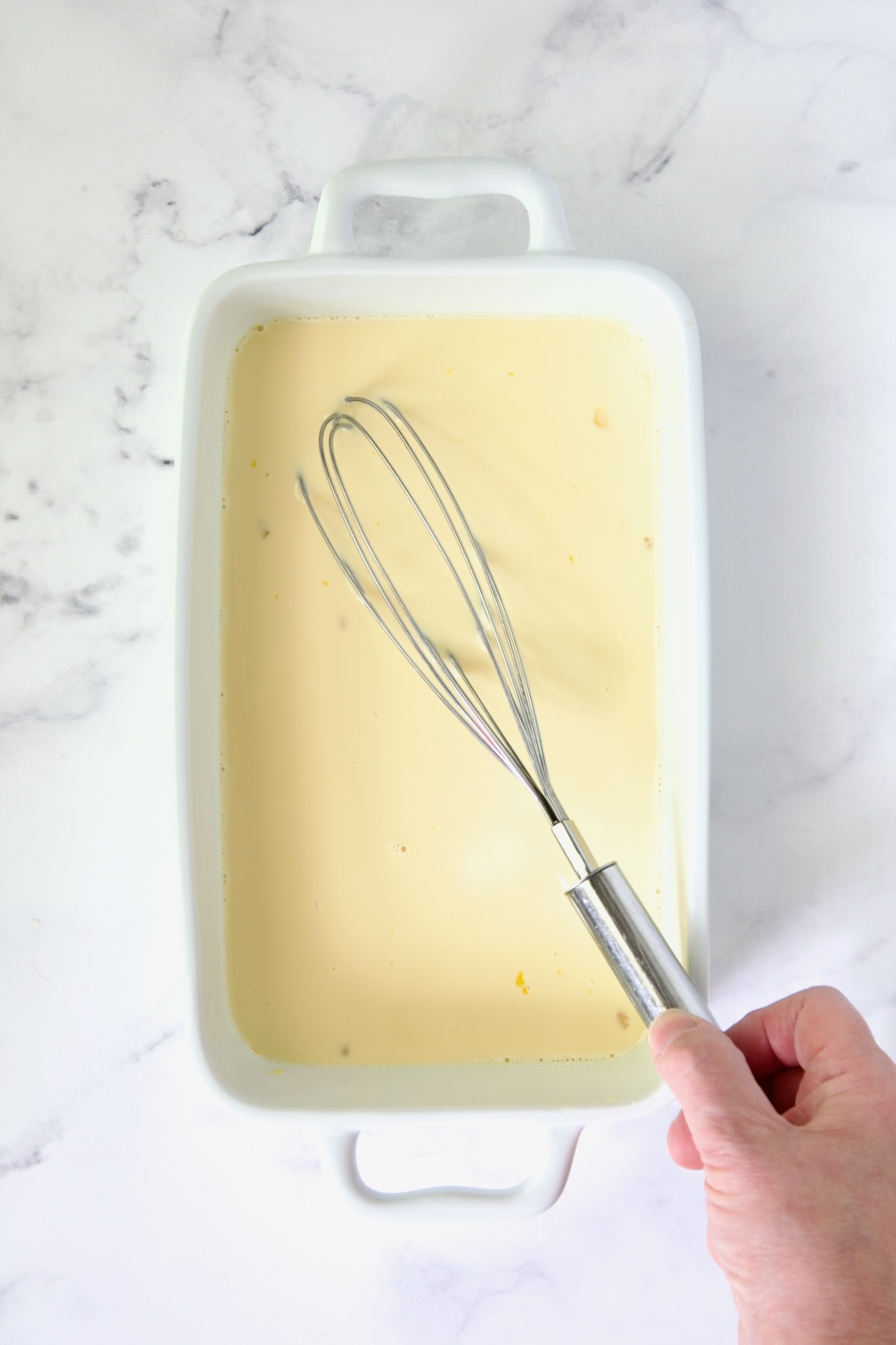 Custard in white rectangular dish being whisked with wire whisk.