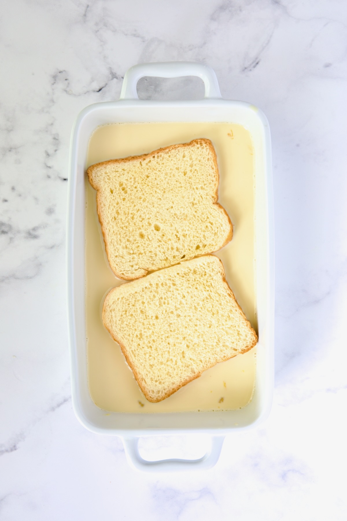 Two slices of brioche bread in custard in white rectangular baking dish before being flipped.