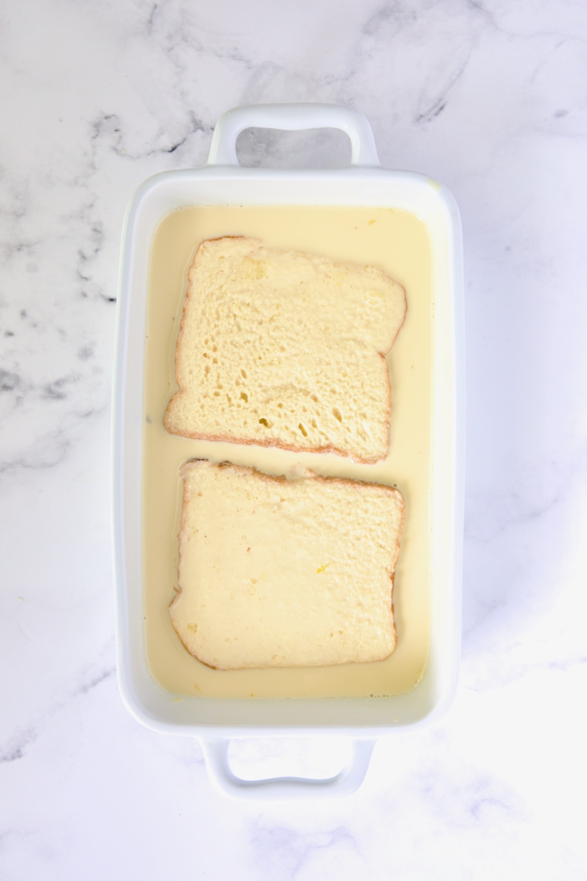 Two slices of brioche bread in custard in white rectangular baking dish after being flipped.