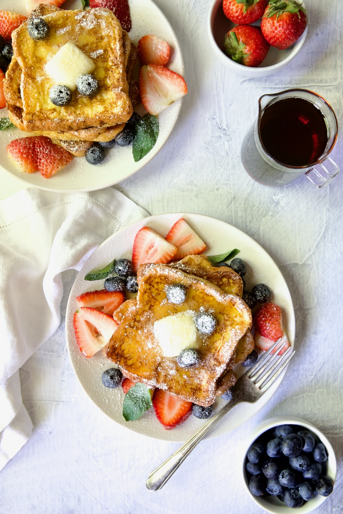 Two plates of Brioche French Toast garnished with fresh fruit, butter pat and powdered sugar.
