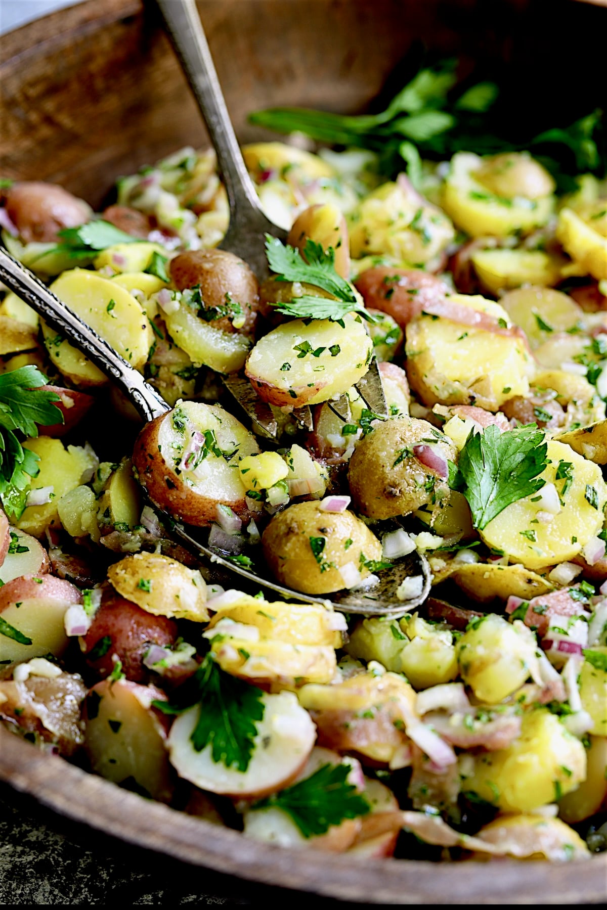 French Potato Salad in wood bowl with serving utensils picking some of it up.