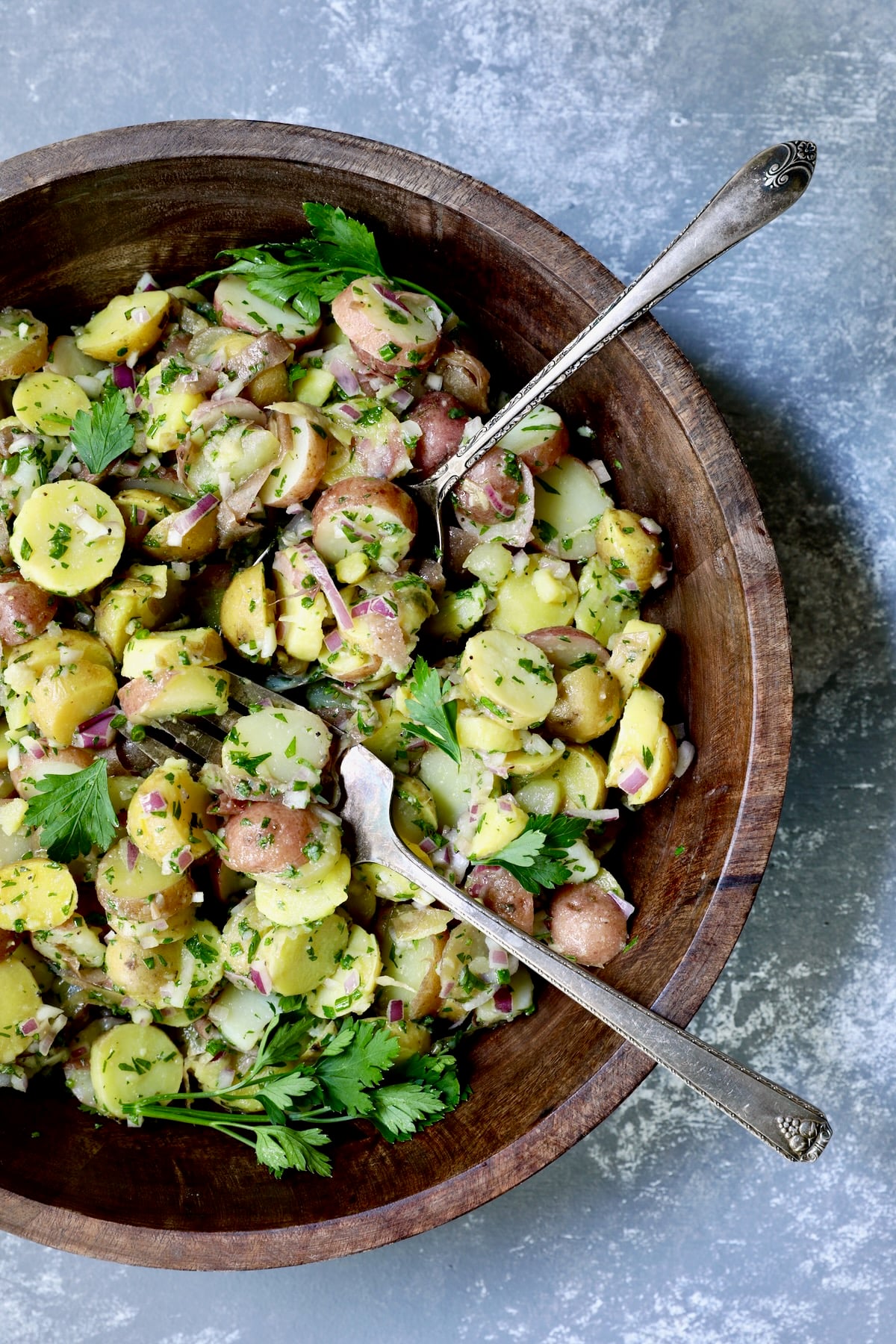 French Potato Salad in wood bowl with serving utensils garnished with fresh parsley.