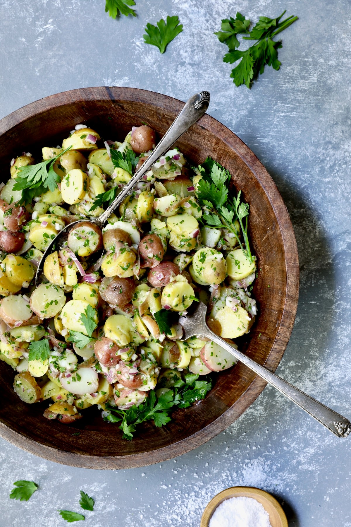 French Potato Salad in wood bowl with serving utensils garnished with fresh parsley with salt scattered about.