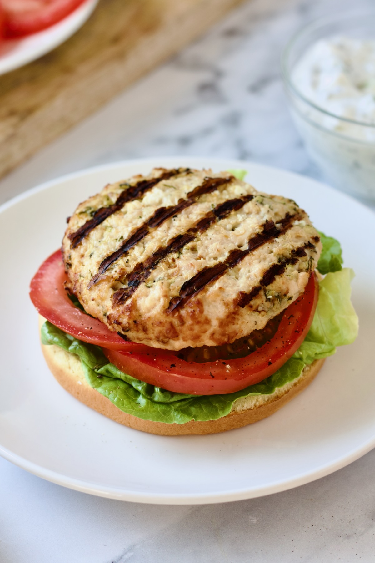 Ground chicken burger with lettuce and tomato on a bun before tzatziki and red onion goes on top.