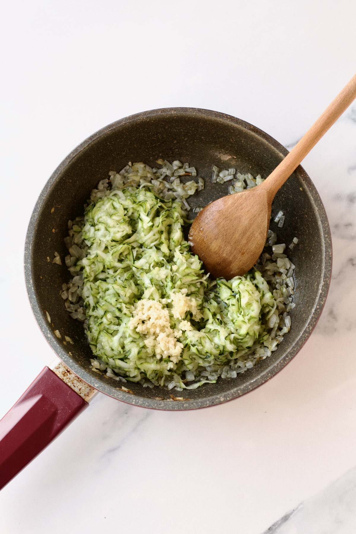 Zucchini and garlic added to sauteed onions in nonstick skillet being stirred with wooden spoon.