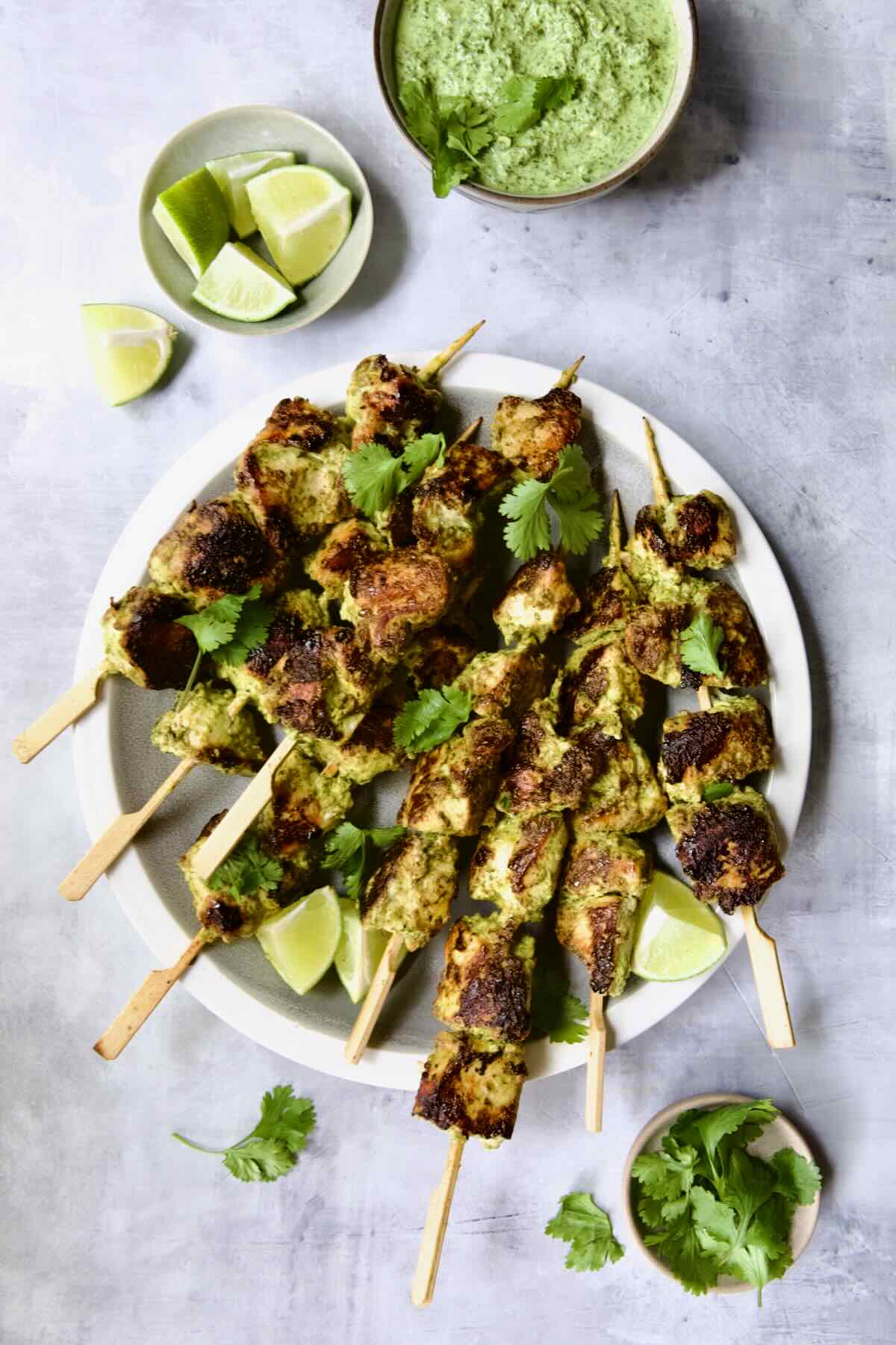 Yogurt Marinated Grilled Chicken Skewers on white plate with Cilantro Mint Chutney, lime wedges and cilantro sprigs in small bowls.