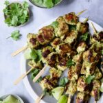 Yogurt Marinated Grilled Chicken Skewers on white plate with Cilantro Mint Chutney, cilantro sprigs and lime wedges in small bowls.