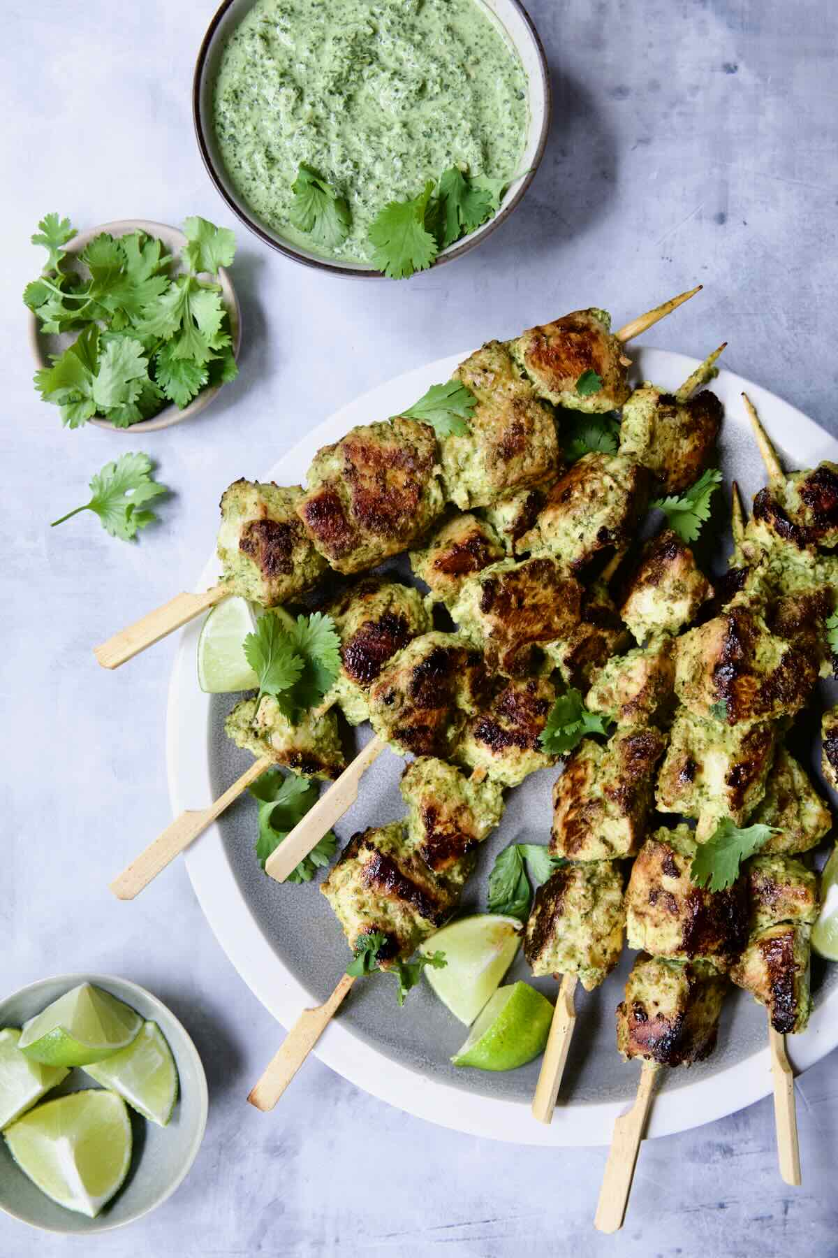 Yogurt Marinated Grilled Chicken Skewers on white plate with Cilantro Mint Chutney, cilantro sprigs and lime wedges in small bowls.