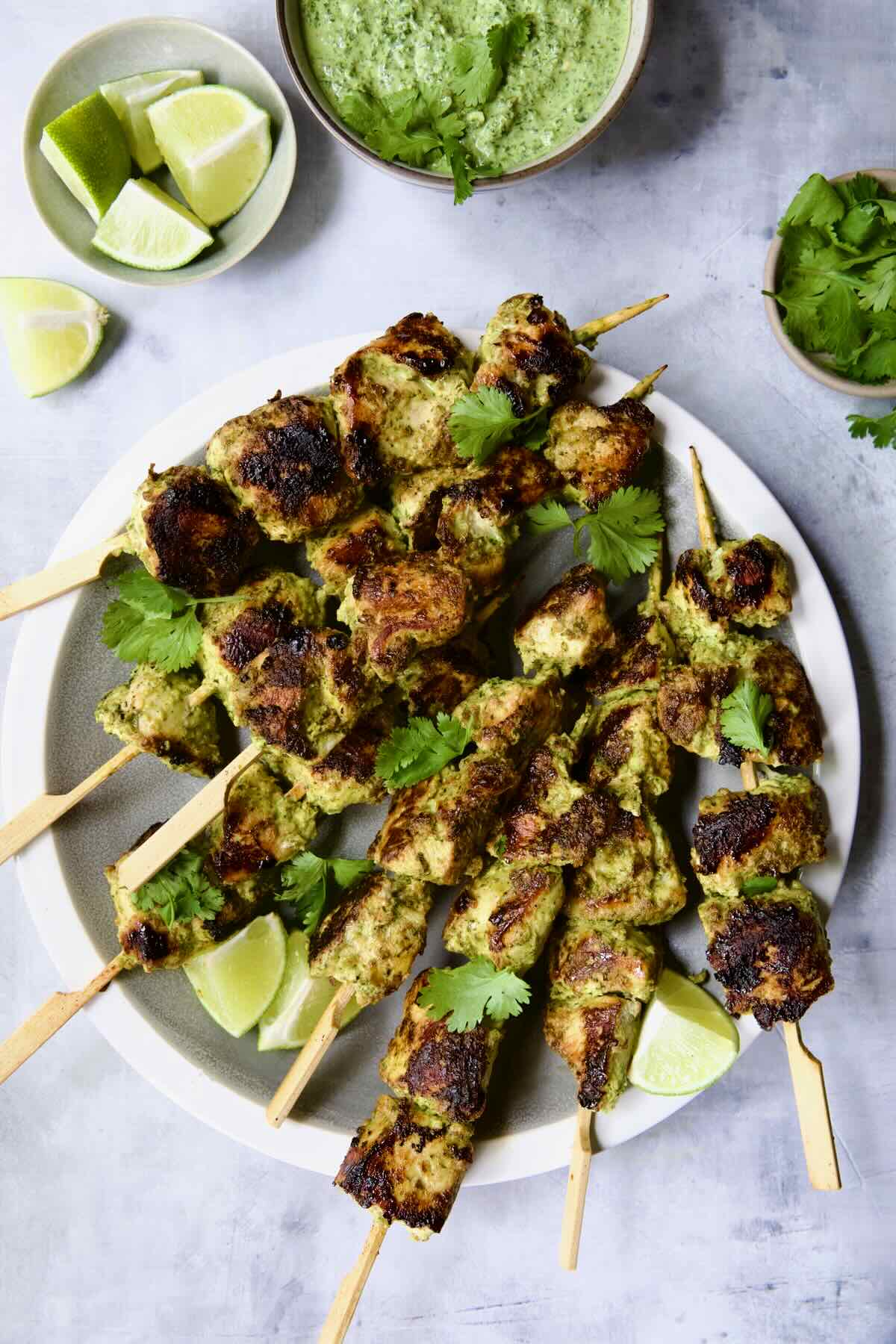 Eight Yogurt Marinated Grilled Chicken Skewers on white plate with Cilantro Mint Chutney and lime wedges in small bowls.