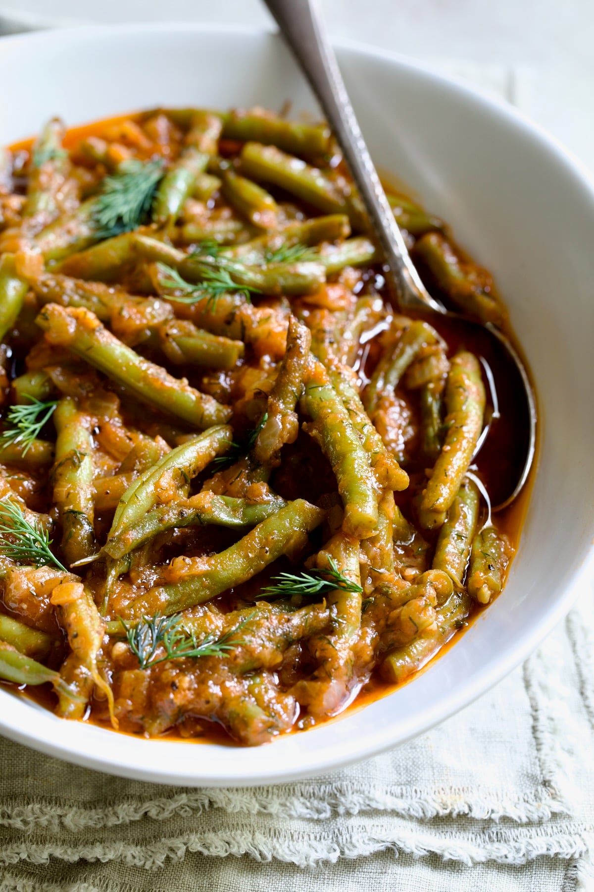 Greek Green Beans in white serving bowl with serving spoon garnished with fresh dill on frayed napkin.