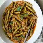 Greek Green Beans in white oval serving bowl with serving spoon on beige frayed napkin.