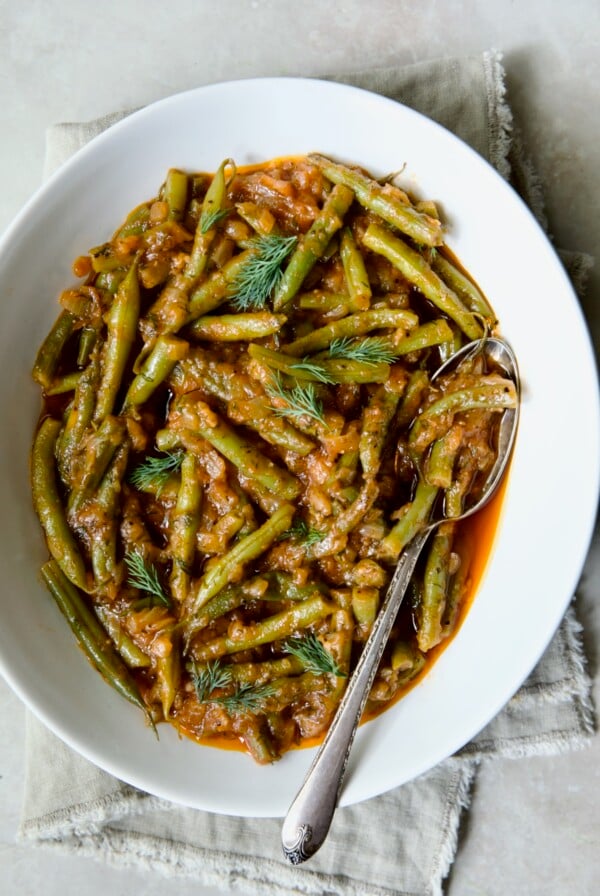 Greek Green Beans in white oval serving bowl with serving spoon on beige frayed napkin.