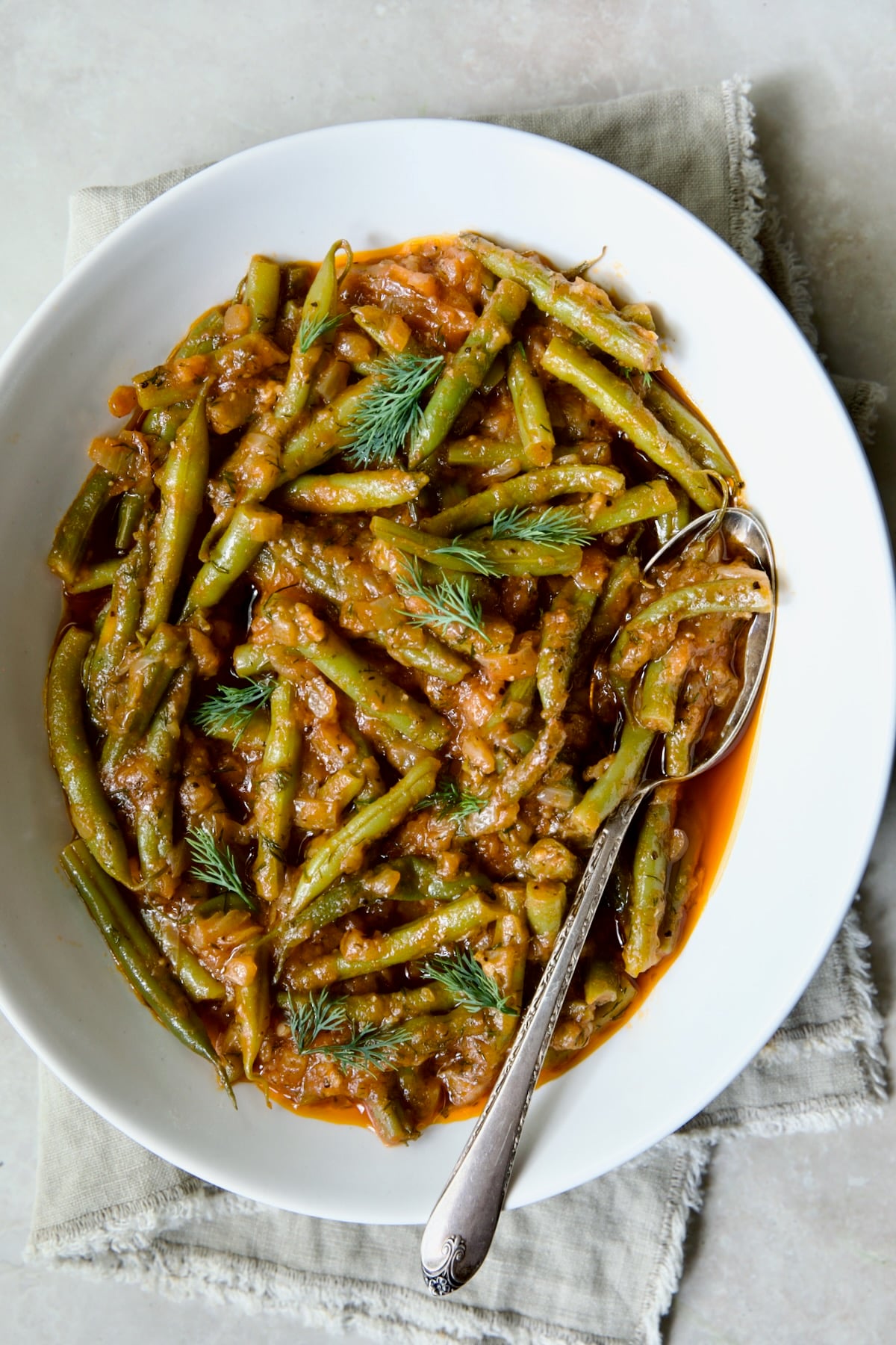 Greek Green Beans in white oval serving bowl with serving spoon on beige frayed napkin.