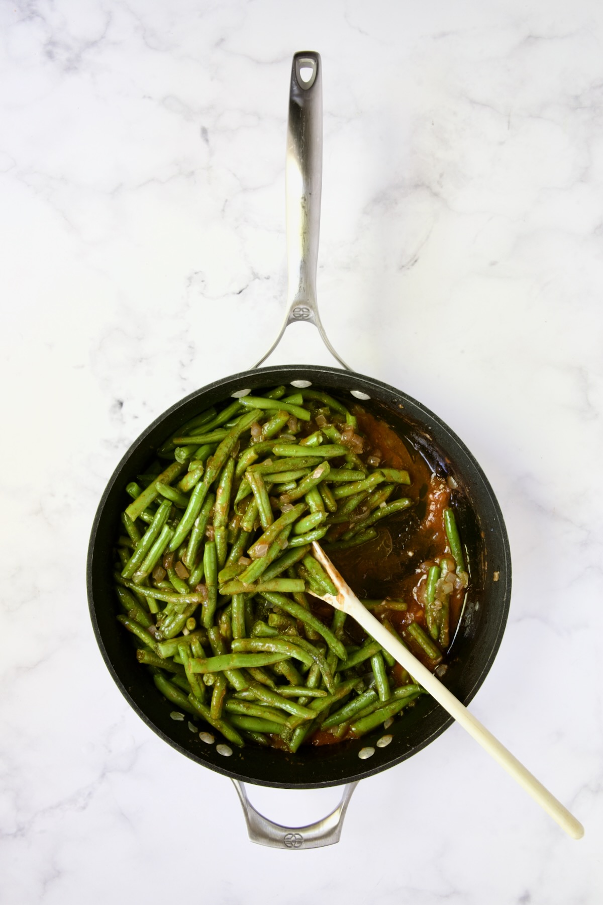 Green beans, tomato sauce and dill being sauteed in nonstick skillet with white spoon.
