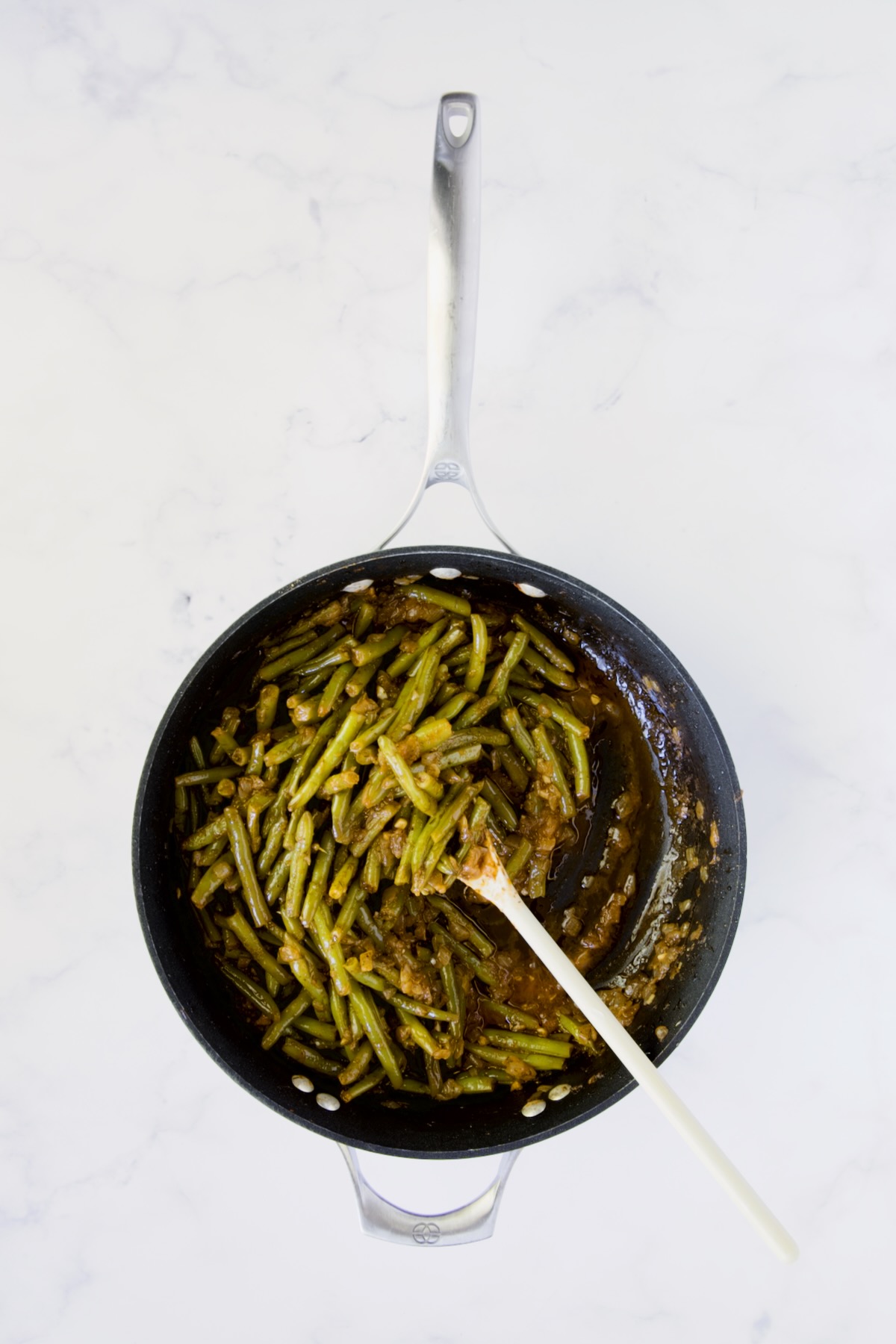 Finished Greek Green Beans in nonstick skillet being stirred with white spoon.