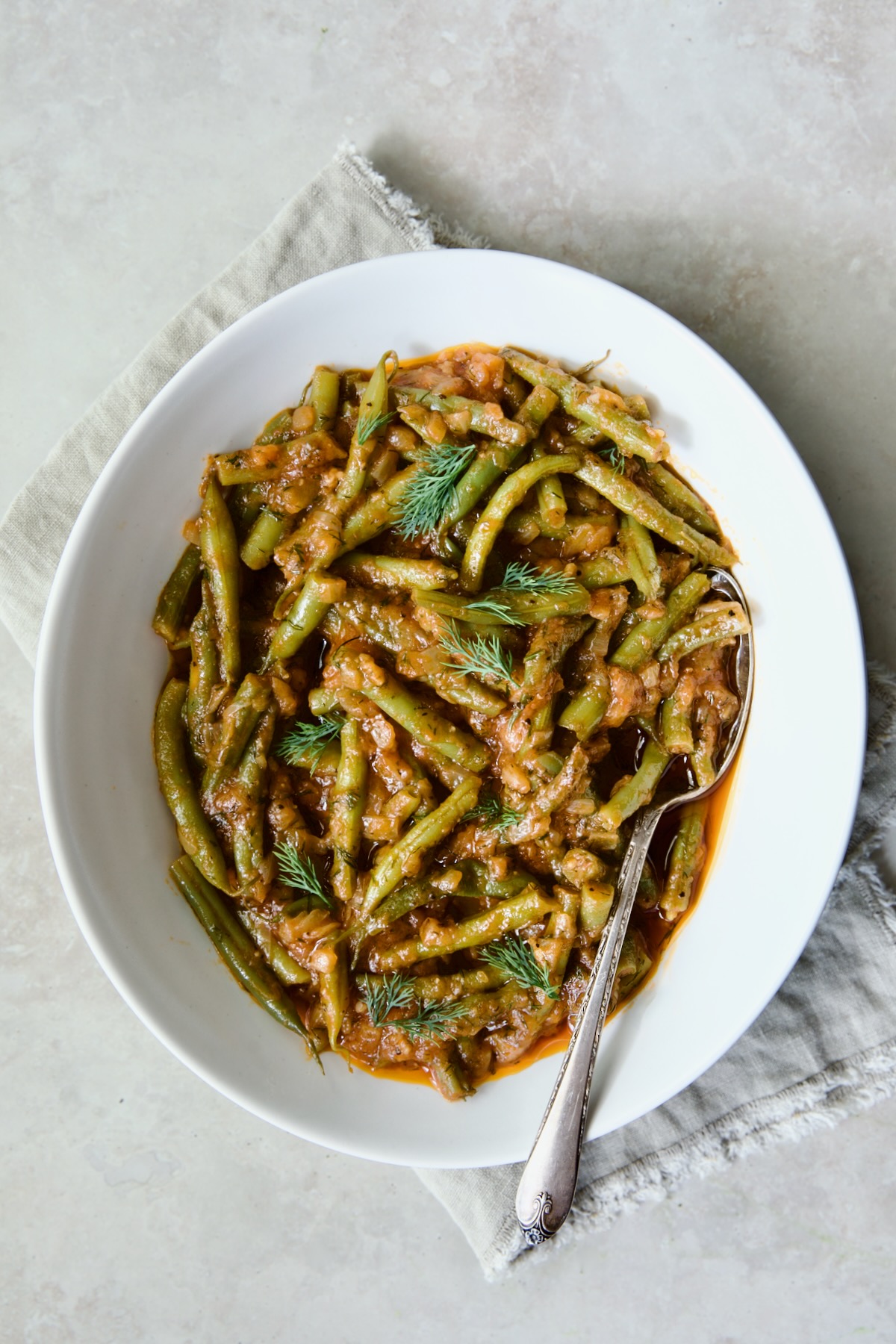 Greek Green Beans in white oval serving bowl with serving spoon on frayed napkin.