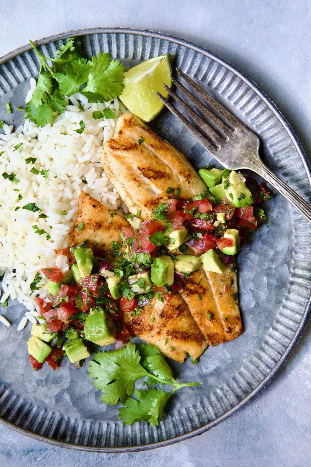 Grilled Tilapia with Tomato - Avocado Salsa on round metal plate with white rice garnished with cilantro and a lime wedge.