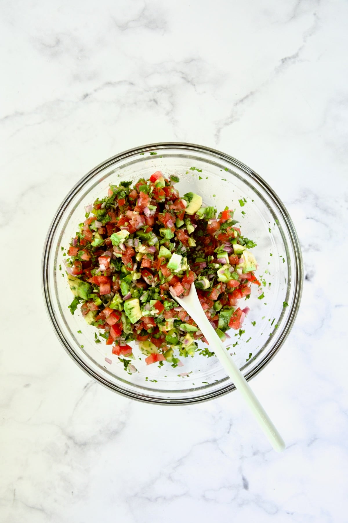 Tomato - Avocado Salsa in glass bowl being stirred with white spoon.