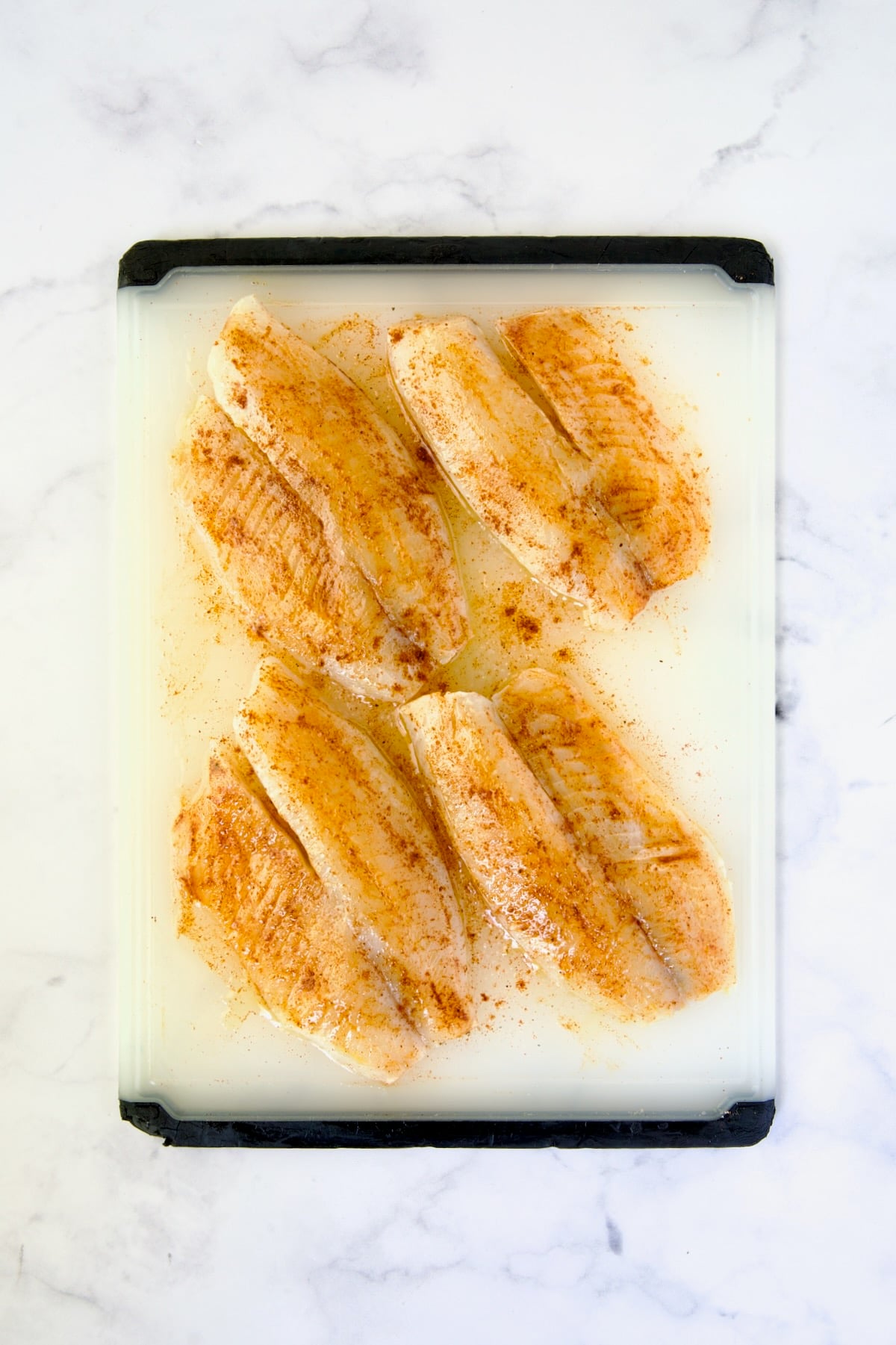 Four pieces of seasoned tilapia fillets on white plastic cutting board.